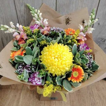 Hand-crafted rustic bouquet by local florist featuring a large yellow chrysanthemum, orange gerberas, snapdragons, and carnations wrapped in bee-patterned kraft paper.