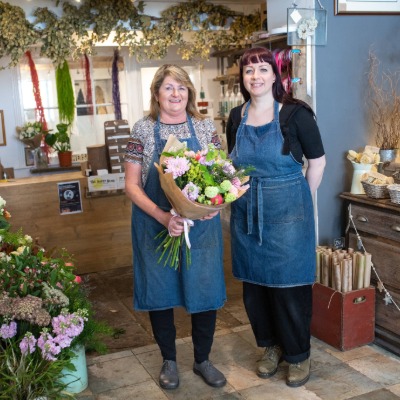 Florists at Flowersmiths in Horwich standing inside their flower shop, holding a colourful hand-tied bouquet surrounded by fresh flowers and floral displays.